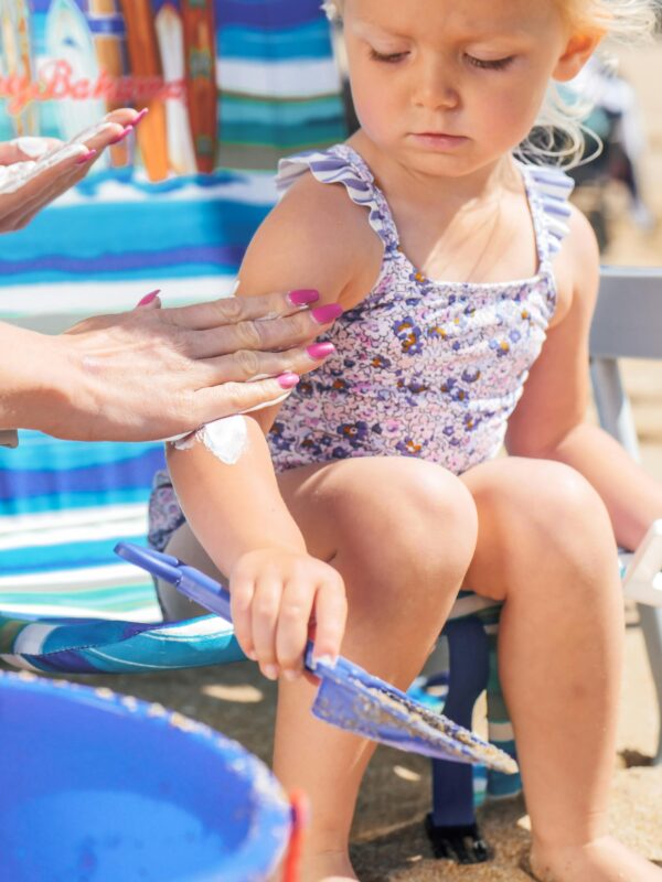 A young girl in a swimsuit playing with sand toys while applying sunscreen on a sunny beach day.